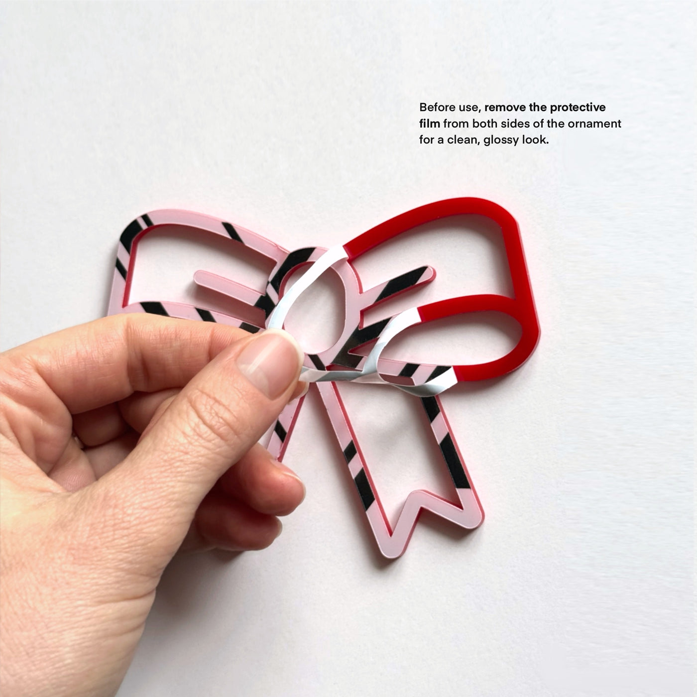 A hand is holding a red and black striped ornament, which appears to be a bow, and is in the process of removing a protective film from both sides of the ornament.