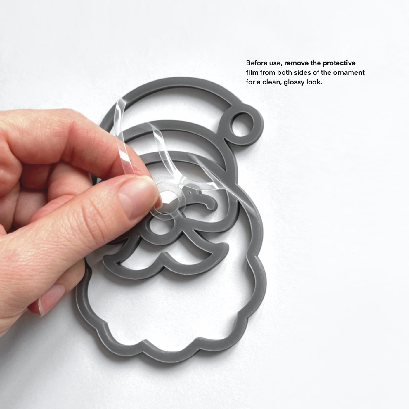 A hand is holding a clear plastic ornament with a Santa Claus shape, and there is text instructing to remove protective film from both sides of the ornament for a clean, glossy look.