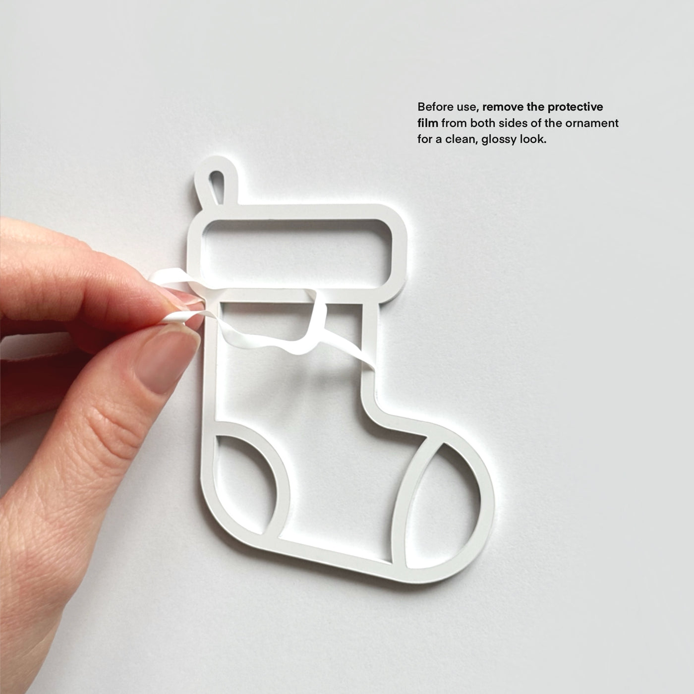 A hand is shown removing a protective film from the sides of a white Christmas stocking ornament.