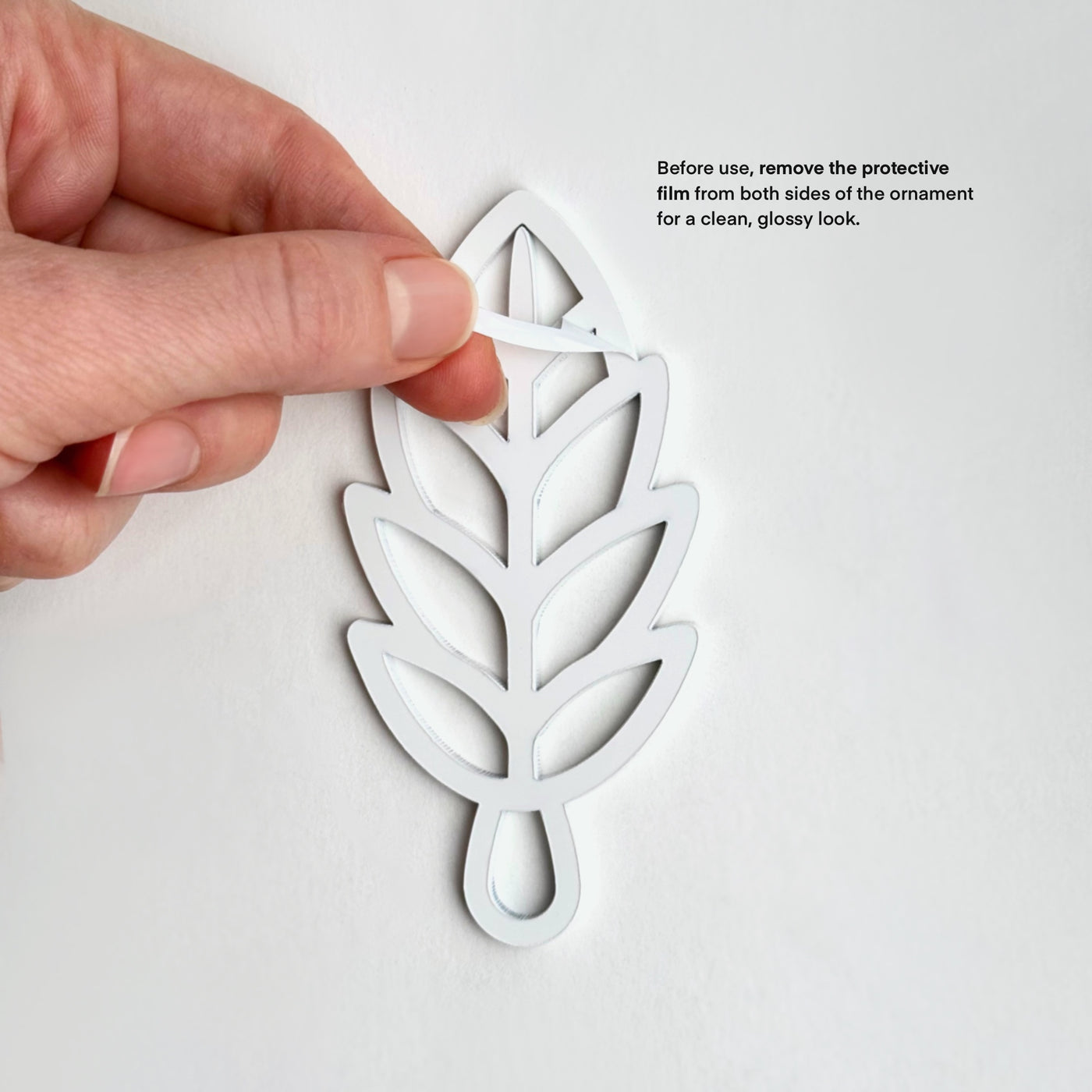 A hand is holding a white leaf-shaped ornament against a white background, with instructions on how to remove the protective film from both sides for a clean, glossy look.