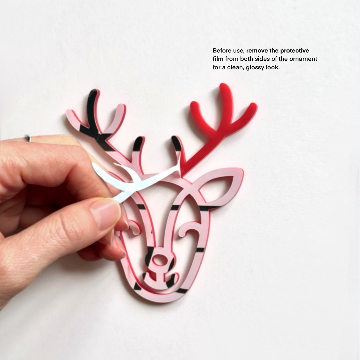 A hand is holding a pink and black ornament resembling a deer head, with the text "Before use, remove the protective film from both sides of the ornament for a clean, glossy look."