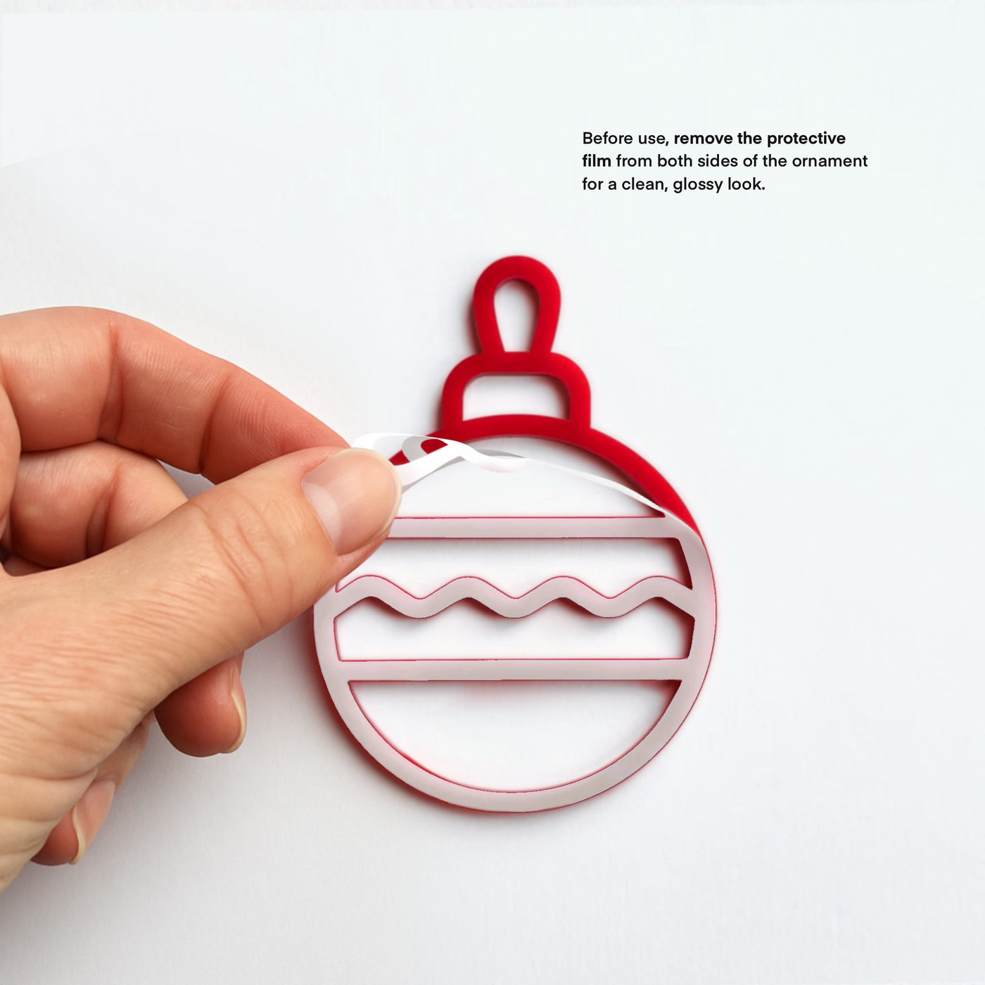 A hand is holding a red ornament with a white and red design, and there is text instructing to remove the protective film from both sides of the ornament for a clean, glossy look.