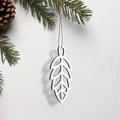 A white ornament resembling a leaf hangs from a string, with pine branches and a pine cone visible in the background. - Photo #2
