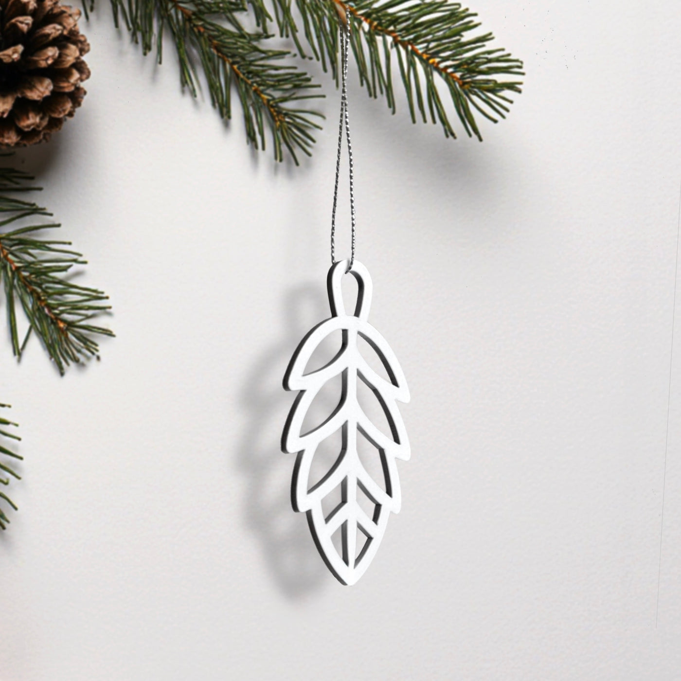 A white ornament resembling a leaf hangs from a string, with pine branches and a pine cone visible in the background.