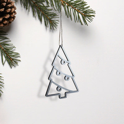 A silver Christmas ornament in the shape of a tree is hanging from a string, with pine branches and a pine cone visible in the background.
