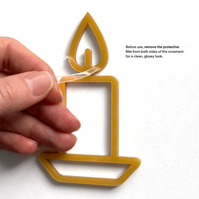 A hand is holding a yellow candle-shaped ornament with a text overlay instructing to remove the protective film from both sides of the ornament for a clean, glossy look.