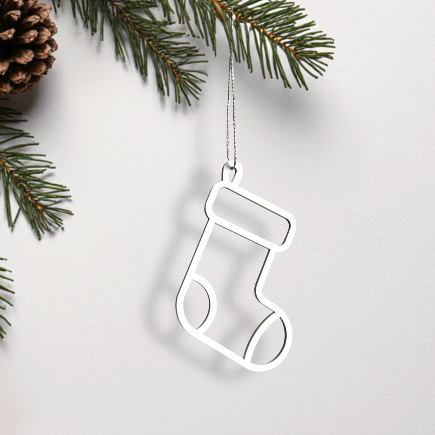 A white Christmas stocking ornament is hanging from a branch, with pine needles and a pine cone visible in the background.