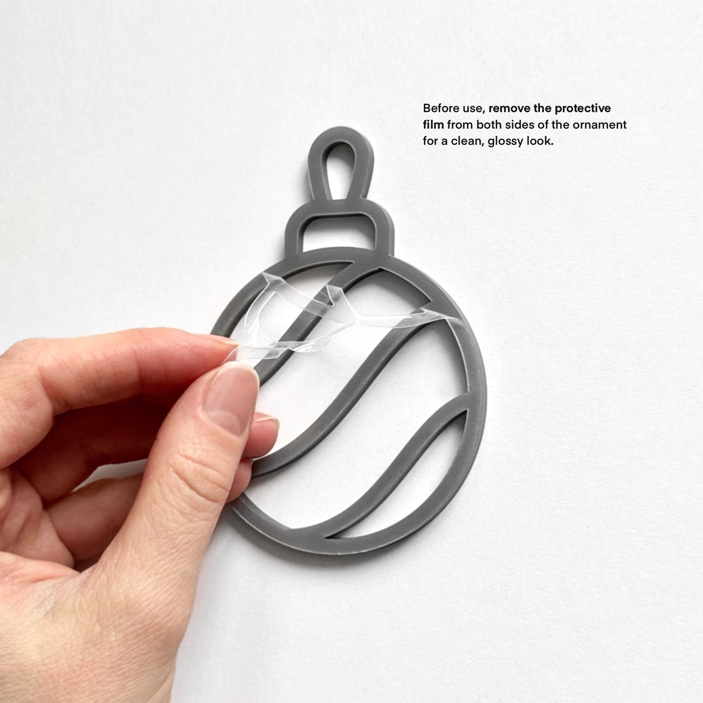 A hand is holding a black ornament with a clear plastic piece attached to it, and there is text instructing to remove the protective film from both sides of the ornament for a clean, glossy look.