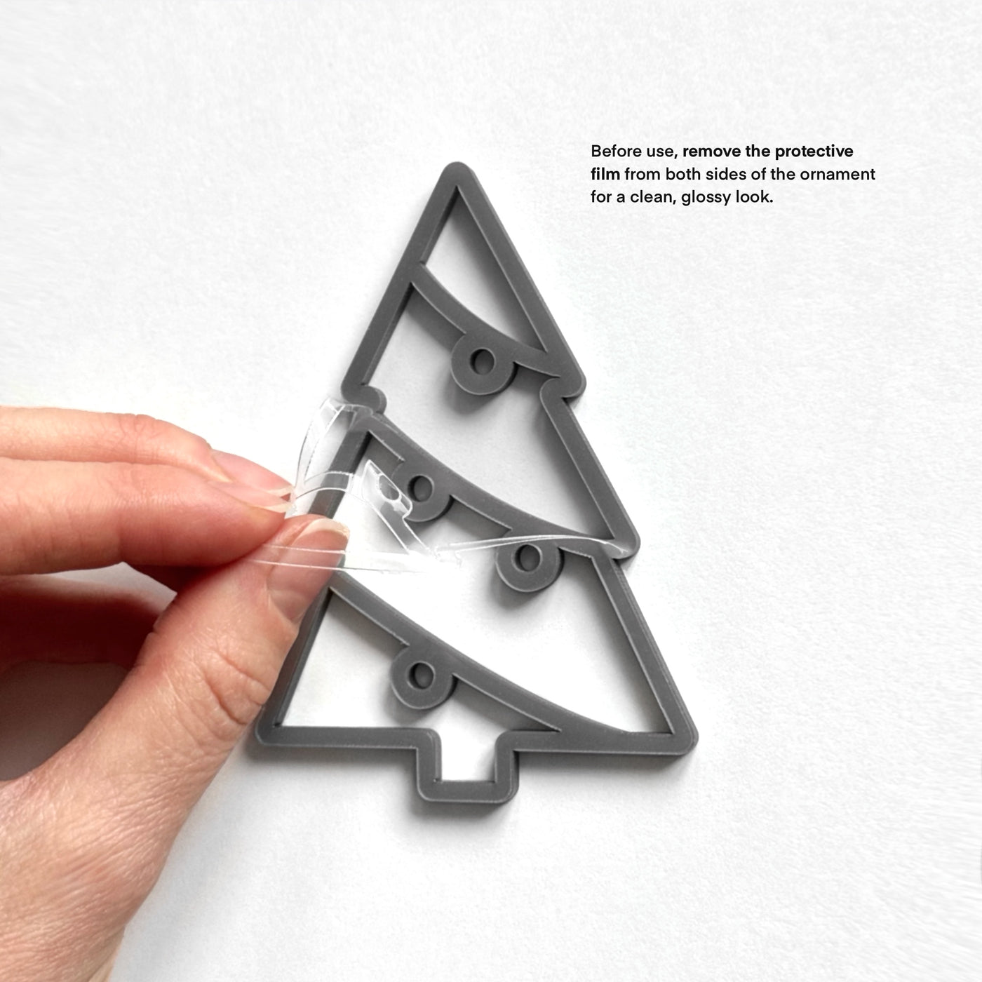 A hand is holding a clear plastic ornament with a Christmas tree design, and there is text instructing to remove protective film from both sides of the ornament for a clean, glossy look.