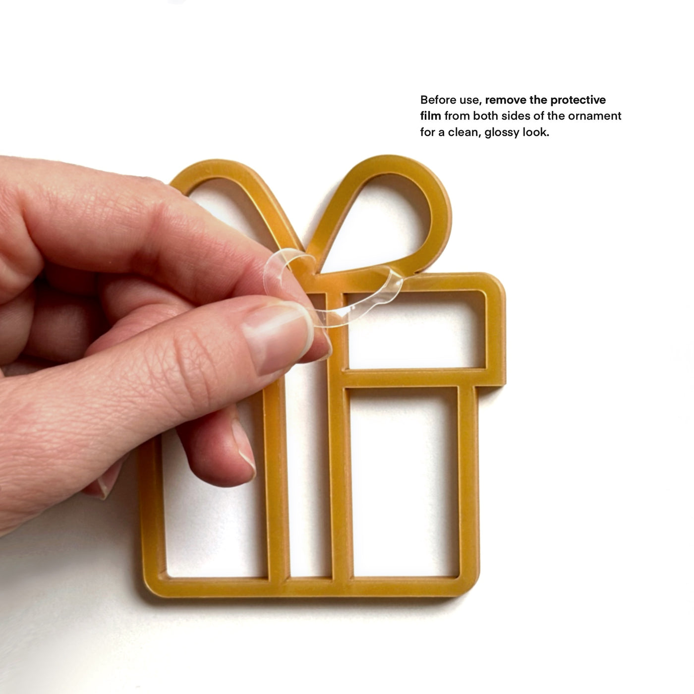 A hand is holding a golden ornament in the shape of a gift box, with instructions to remove the protective film from both sides for a clean, glossy look.