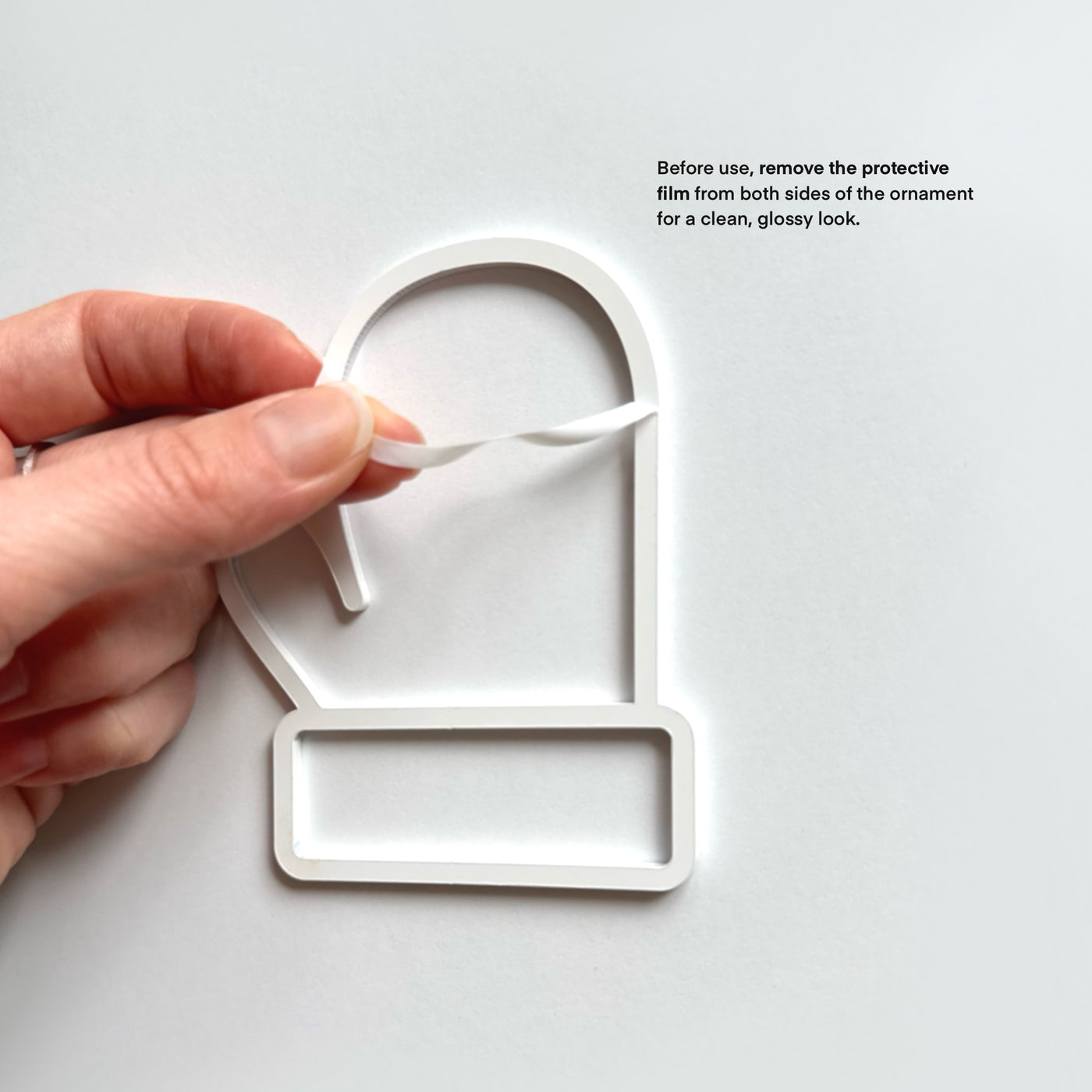 A hand is holding a white ornament with a protective film on both sides, preparing to remove the film for a clean, glossy look.